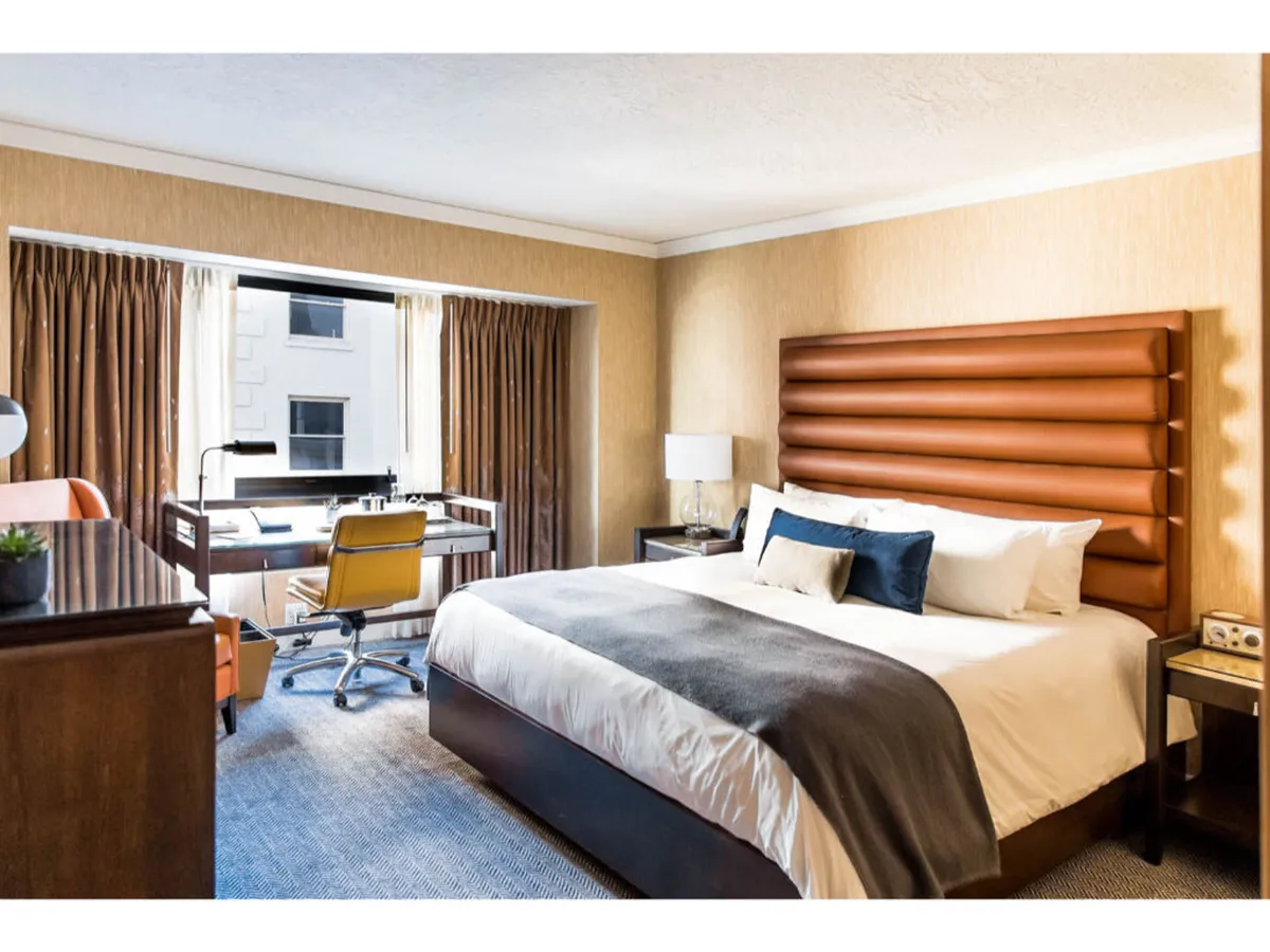 The image shows a stylish hotel room with a plush bed featuring a designer headboard, a sleek work desk and chair, and large windows draped with curtains that let in abundant natural light. The décor combines earthy tones with touches of luxury like velvet and leather, creating a warm, inviting ambiance. The setting suggests both relaxation and productivity, appealing to leisure and business travelers alike.