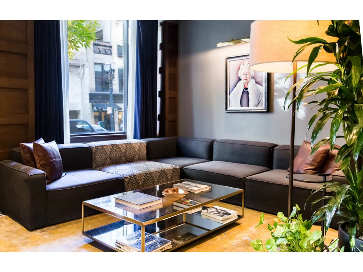 A stylish living area featuring a modern sectional sofa, glass coffee table, framed original artwork, large windows with natural light, and an indoor plant. The space feels airy, welcoming, and artfully designed.