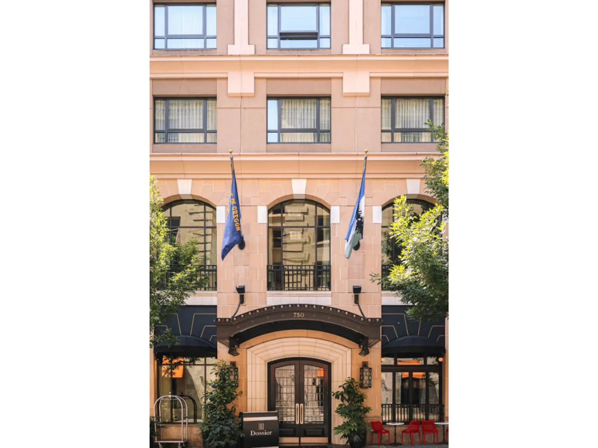 The main entrance to the hotel, framed by stylish landscaping, large windows, and flags. Guests can be seen arriving or socializing in the outdoor area, giving a sense of arrival and urban connection.