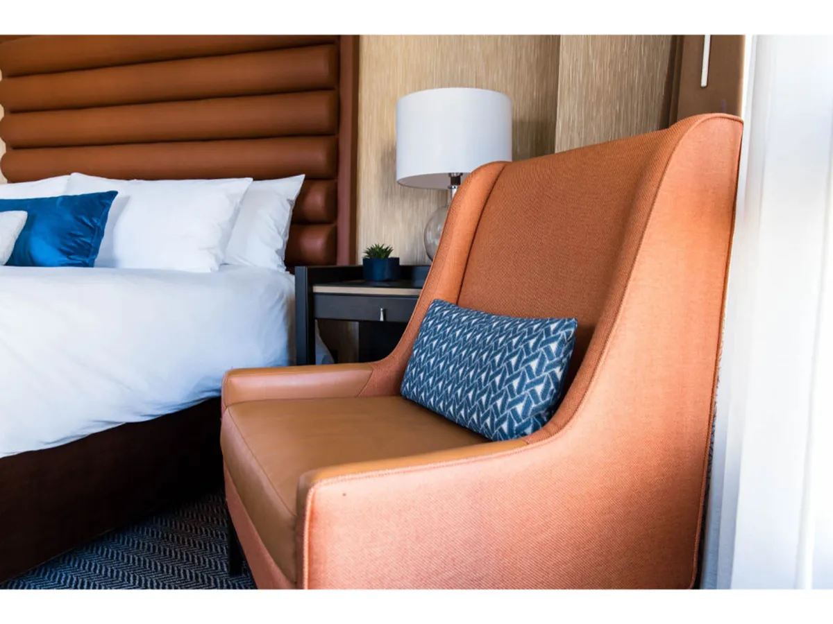 A cozy guest room featuring a plush bed with crisp white linens, an orange accent chair, and a blue decorative pillow. The setting suggests guests can relax with a book or enjoy morning coffee in a bright, comfortable space.