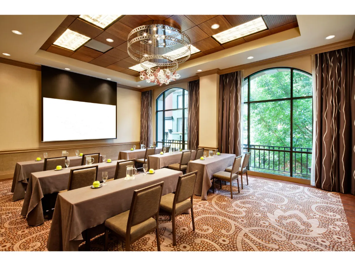 A spacious, well-lit meeting space featuring long tables arranged for a professional gathering. The room is filled with natural light streaming in from large windows, with a classic chandelier overhead adding a touch of sophistication. The layout supports collaborative work and focused business activities.