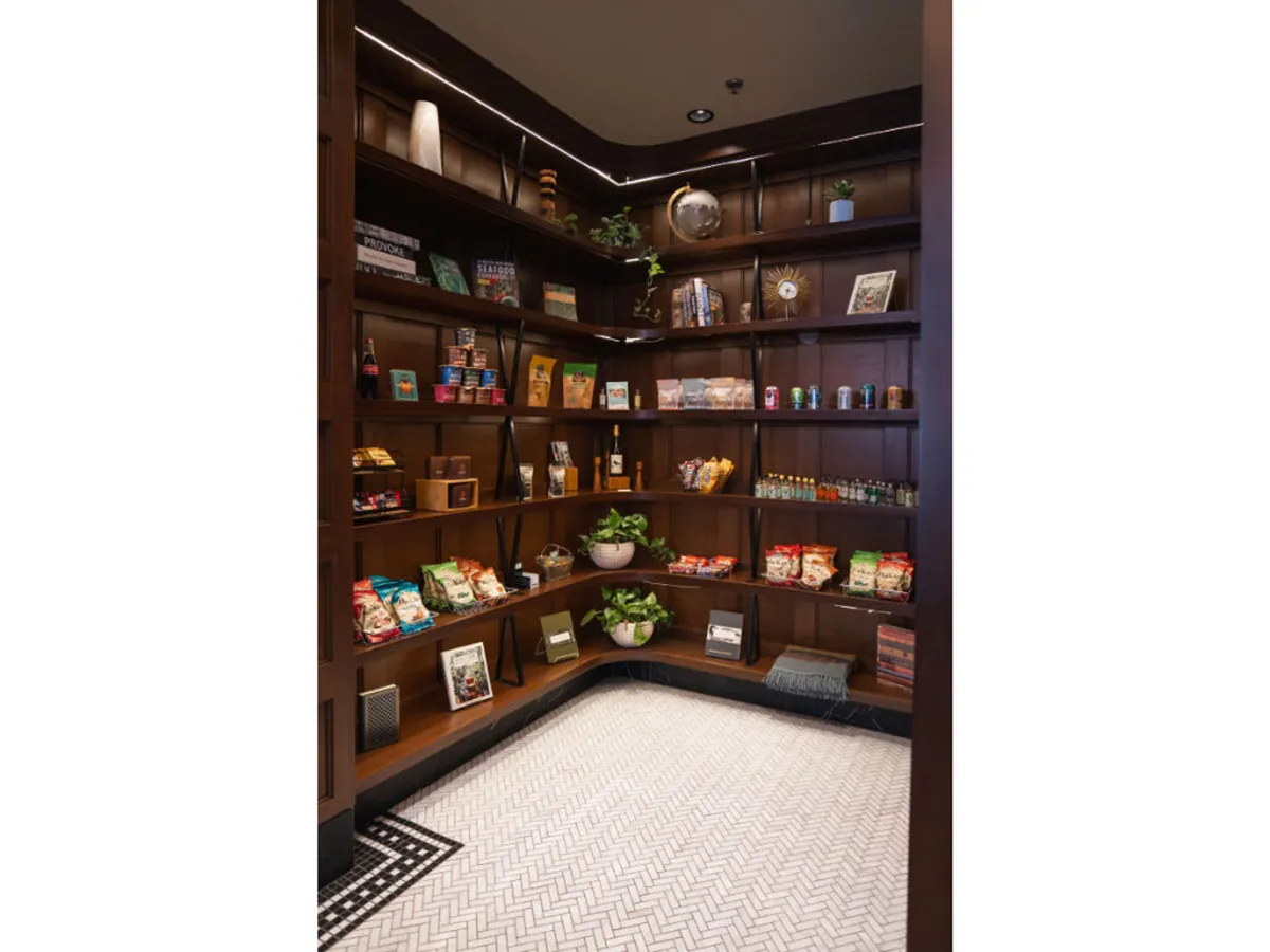 A casual lounge space with wooden shelves stocked with snacks and beverages, decorative plants, and inviting patterned flooring. Guests are seen browsing and socializing, creating a welcoming and home-like environment.