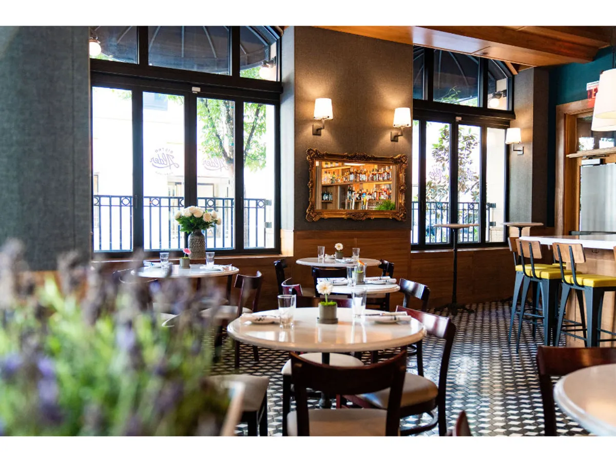 A sunlit dining area with large windows, elegantly set tables, and a decorative mirror reflecting the bar in the background. Lavender plants in the foreground add a cheerful touch, while guests enjoy casual meals and friendly toasts, underscoring the space’s relaxed, welcoming energy.