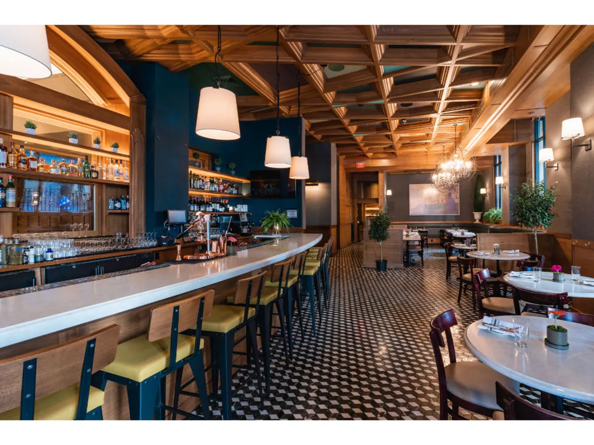 A wide view of Bar Alder’s long bar with stools, featuring ornate ceiling details, elegantly arranged bar shelves with bottles, and adjacent dining tables. The space is filled with guests sharing small plates and unique drinks, emphasizing the culinary diversity and social energy of the venue.