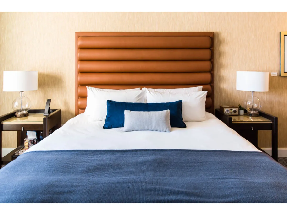 This image highlights a tranquil bedroom with a plush king-sized bed featuring an upholstered headboard, crisp bedding, decorative pillows, and soft lighting from nightstand lamps. The textures and cozy design convey comfort and relaxation.