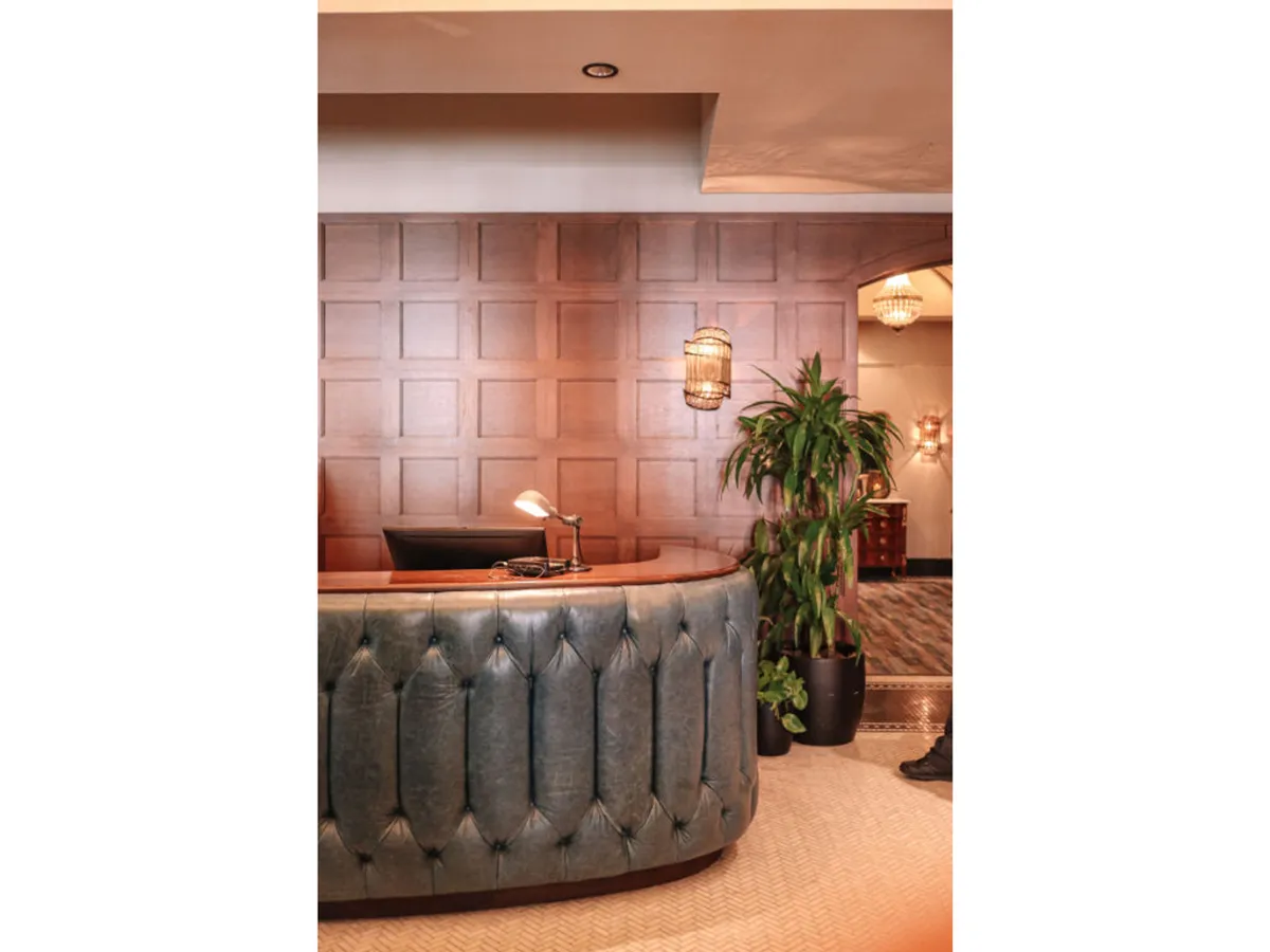 A modern, curved reception desk set against warm wood paneling, complemented by a potted plant and stylish hanging lights. A hotel staff member is visible, engaging with a guest in a friendly manner. The inviting atmosphere sets the tone for attentive service.