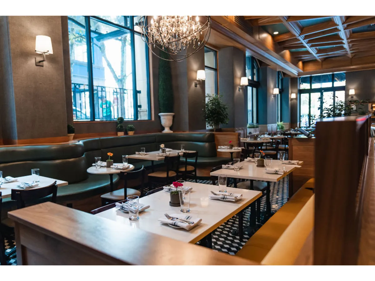 This image features a softly lit dining room adorned with a striking chandelier, lush green seating, and decorative plants. Natural light streams in from a large window, illuminating the intimate arrangement of tables—each set for guests and suggesting an inviting atmosphere for elegant meals or quiet moments together.