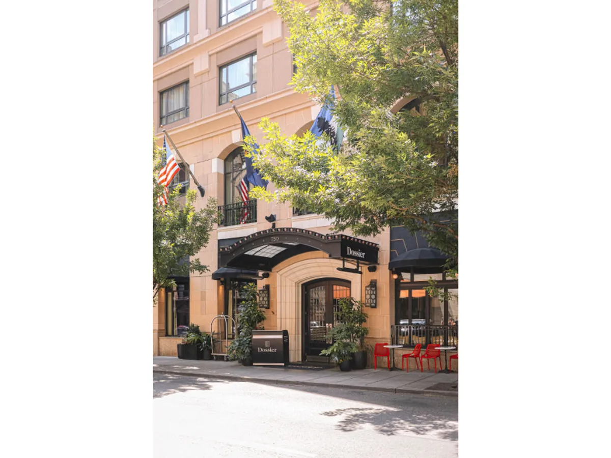 A welcoming hotel entrance with an awning, fluttering flags, potted plants, and inviting outdoor seating. The view captures the energy of the surrounding city and hints at the lively atmosphere just steps away.